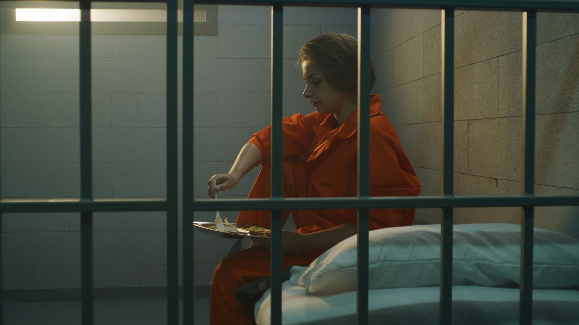 A woman in an orange jumpsuit sits behind prison bars sitting on a bed and ...