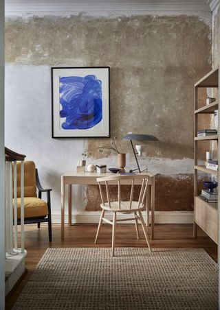 home office with pale blonde wood desk and chair, rough textured plaster wall, blue artwork, storage unit, armchair, textured rug in neutrals