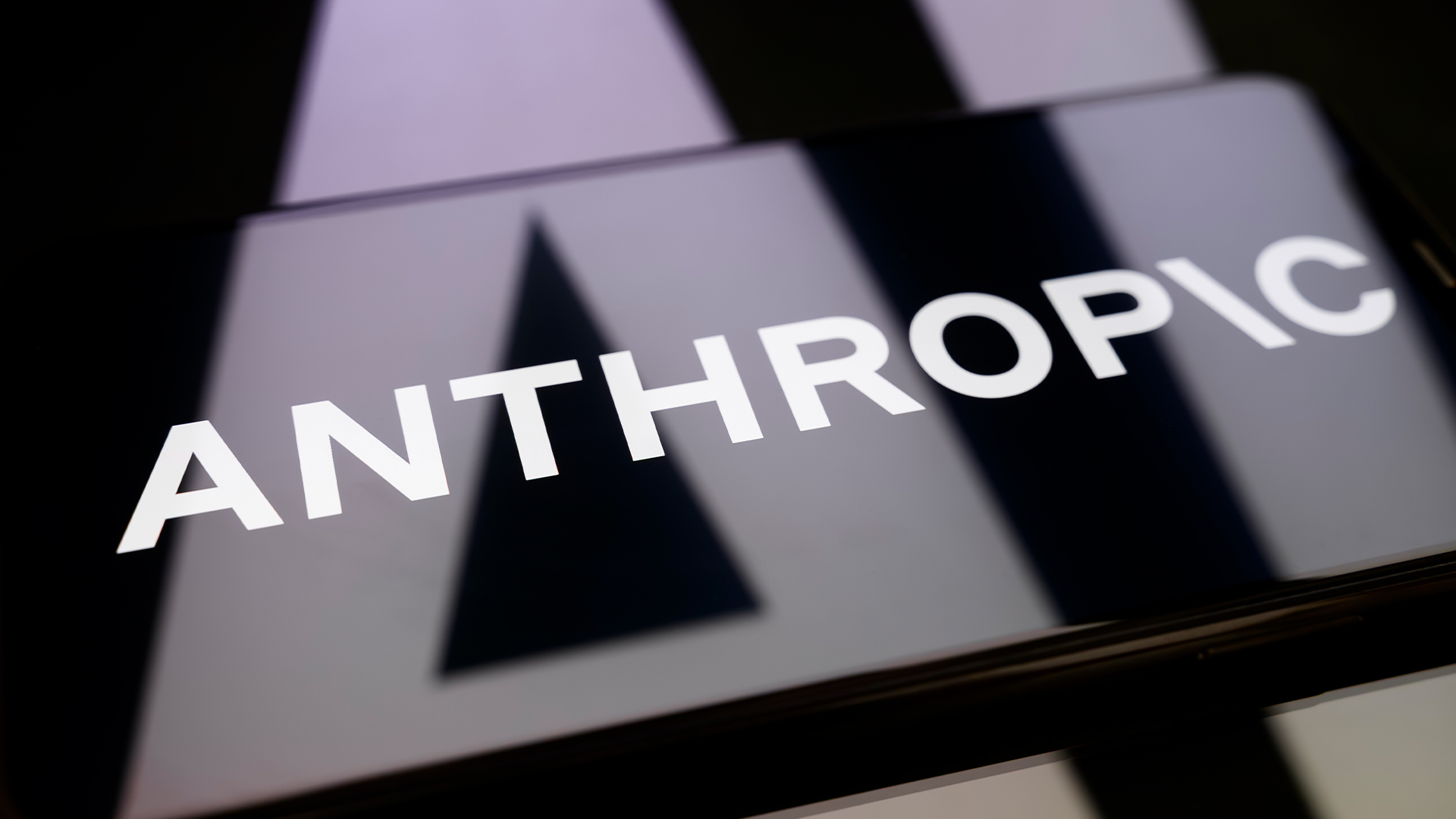 Logo of AI company Anthropic pictured on a smartphone screen placed on top of reflective surface, with company logo reflected.
