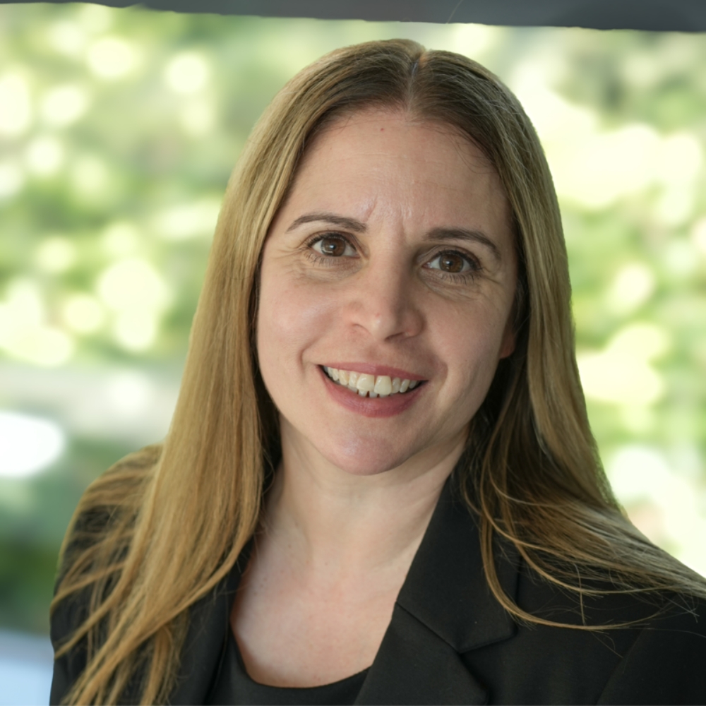 Smiling woman with long straight blonde hair, wearing a black blazer, headshot with a leafy blurred background.