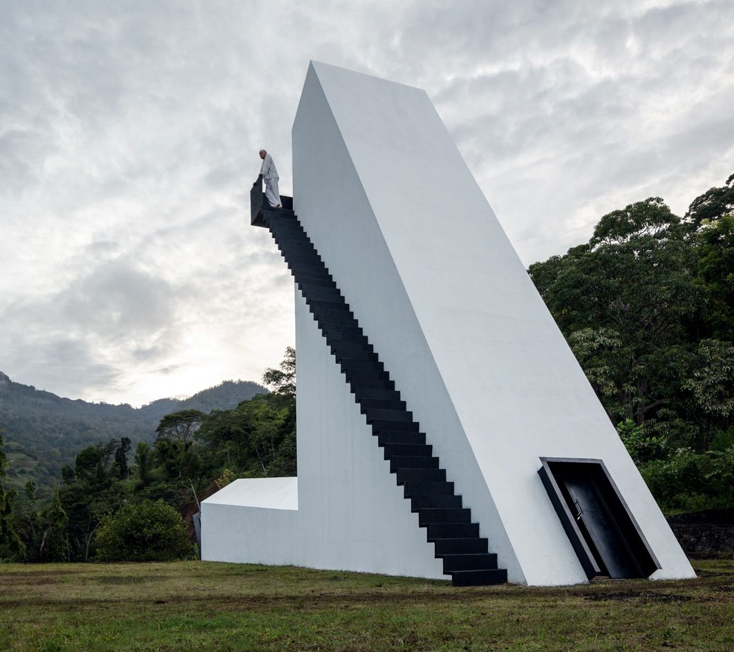 A slant shaped white residential home featuring a black front door and black staircase on the left of the room which leads from the ground to the roof top with views of the forest