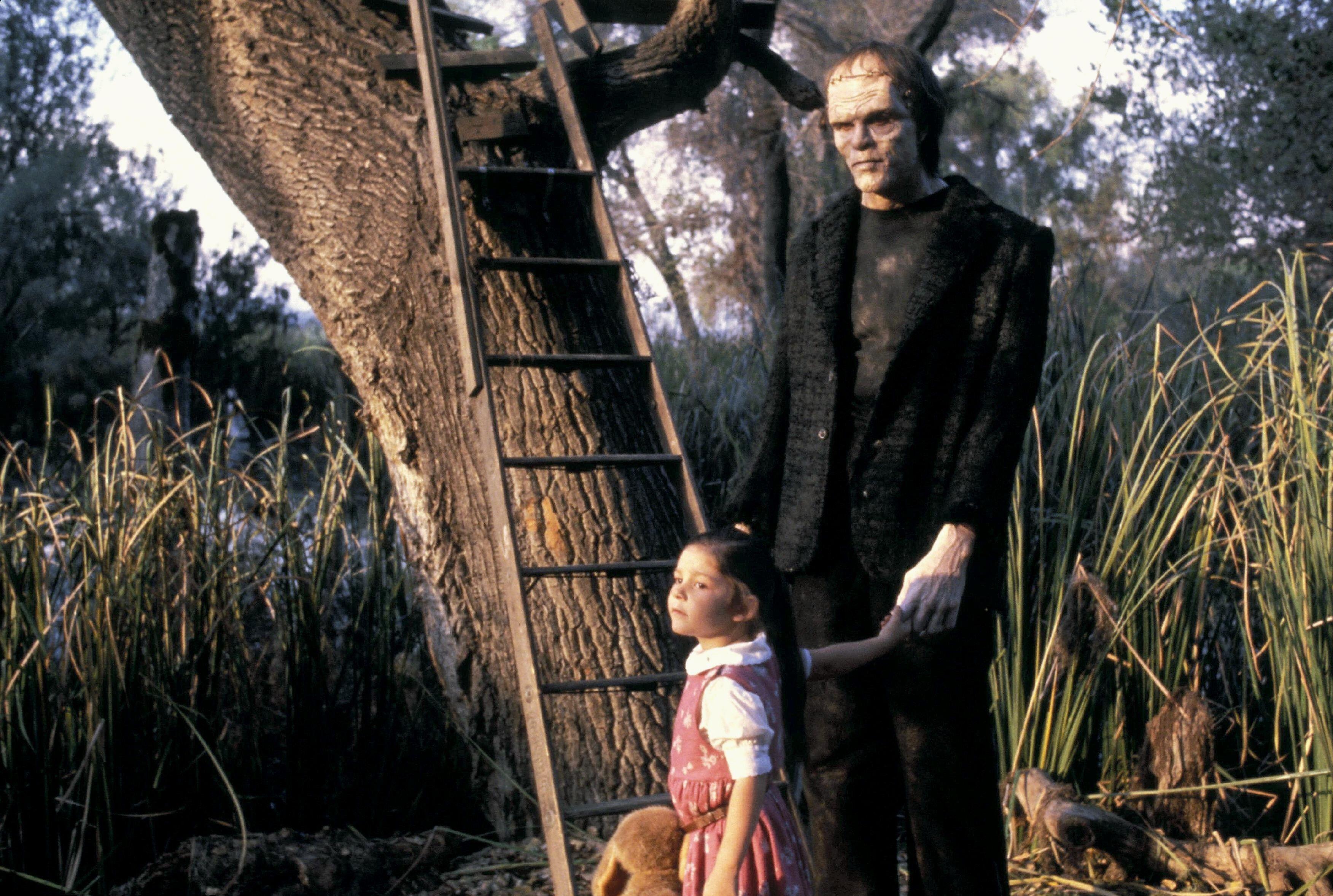 frankenstein stands next to a small girl by a tree in a still from the movie the monster squad 1987