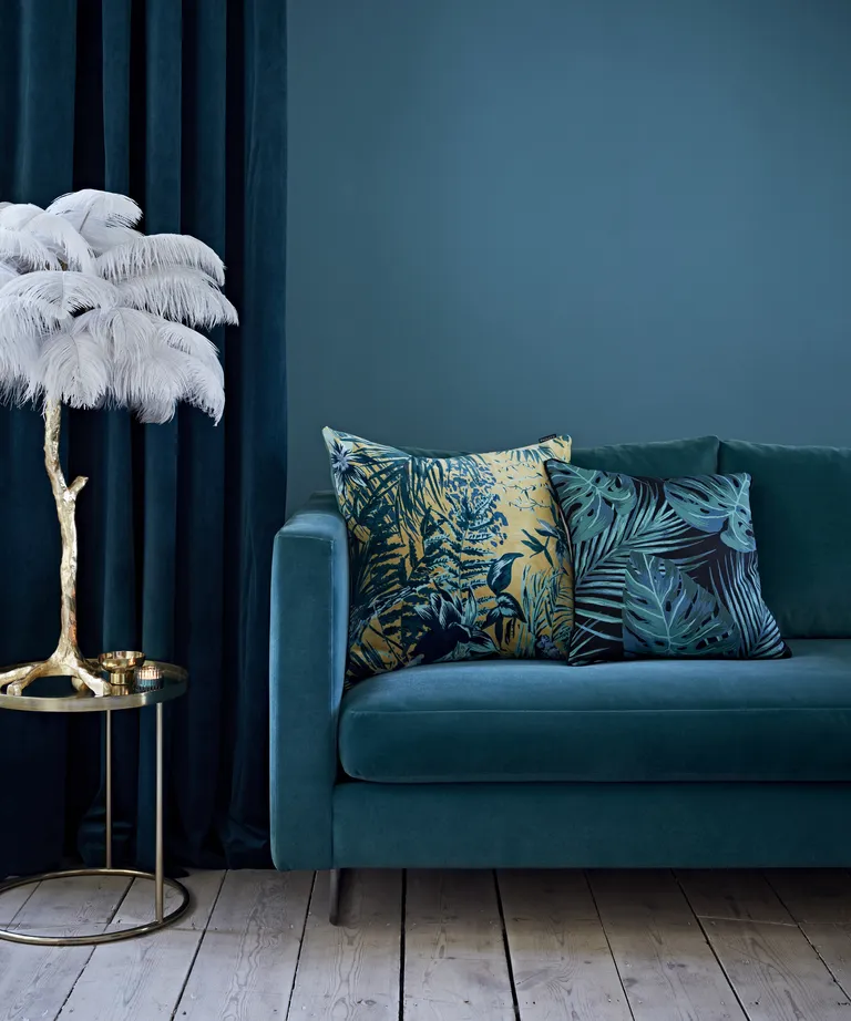 A living room with dark turquoise walls, blue velvet sofa and curatins and a gold side table with feather ornament