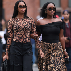  A guest (L) wears Miu Miu brown sunglasses, gold pendant chain necklace, brown animal print / leopard print sheer long sleeve top, black cargo long pants with pockets and Bottega Veneta black woven leather pouch / clutch bag; Next to her a guest (R) seen wearing Celine brown oval Triomphe sunglasses, Dior vintage gold earrings, diamond pendant necklace, black cotton off-shoulder top, brown animal print / leopard print flared pleated long skirt and Bottega Veneta black woven leather mini Jodie bag, outside the Alice + Olivia by Stacey Bendet Spring 2025 presentation, during the New York Fashion Week, on September 07, 2024 in New York City. 