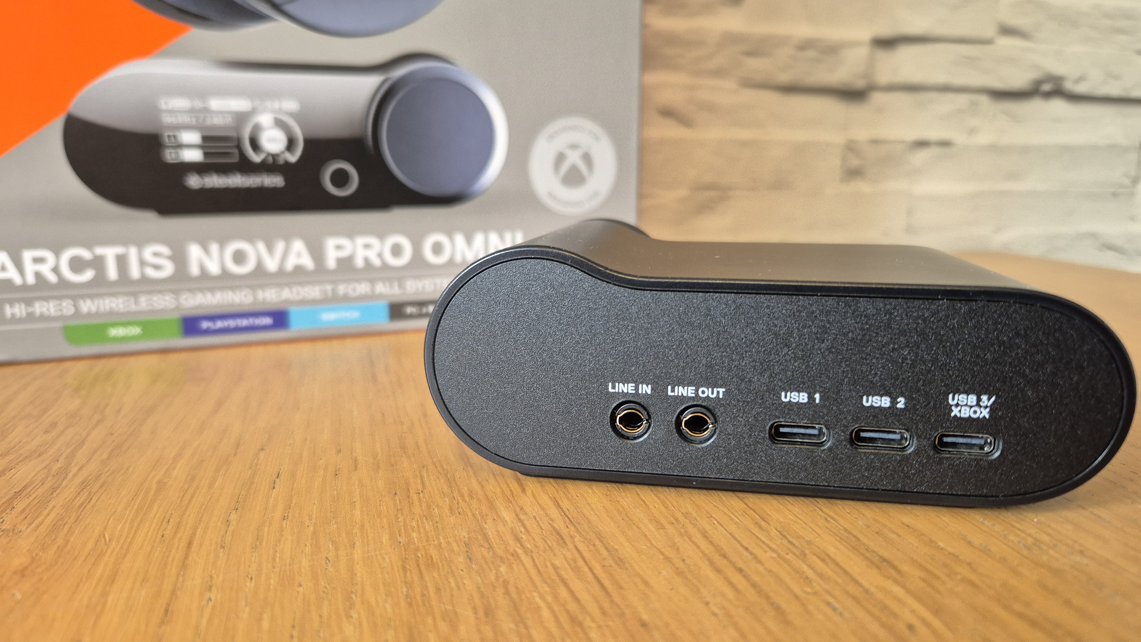 The SteelSeries Arctis Nova Pro Omni's GameHub in front of the product box and a white brick background and on a wooden surface.