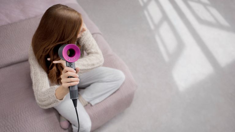 A woman sitting on the edge of her bed, drying her hair with the best hair dryer – the Dyson Supersonic