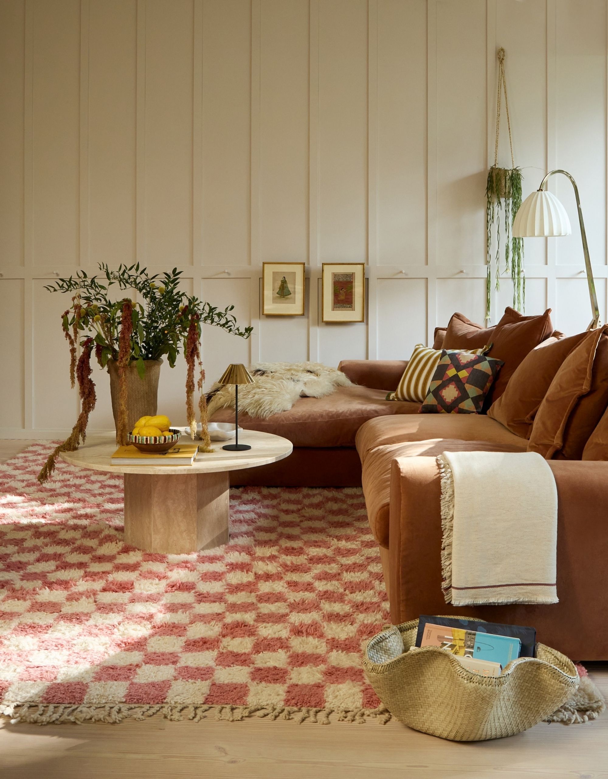 Image of a white living room with a fluffy pink and white checkerboard rug, a brown sofa, and a round coffee table that has a plant and a lamp on it. There is also a floor lamp.