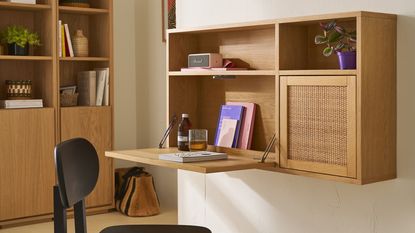 bookshelf in background with bag on floor beside it, black chair in front of cane-fronted wall cabinet that folds down as a desk with books, water glass, speaker, and potted plant
