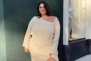 Plus size influencer Gia Sintra wearing off-white asymmetrical knit dress with white thigh-high boots, standing in front of a greeen wall.