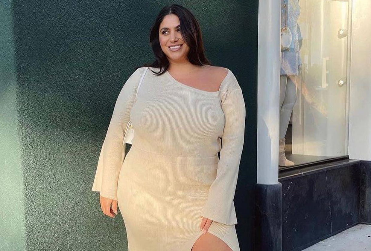Plus size influencer Gia Sintra wearing off-white asymmetrical knit dress with white thigh-high boots, standing in front of a greeen wall.