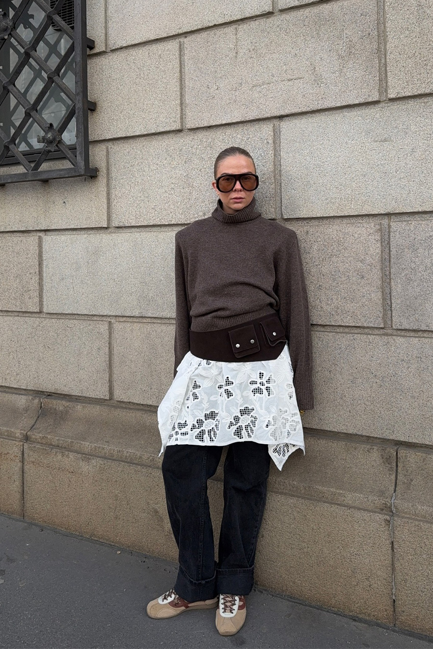 A sweater styling trend is shown in a photo of a woman&#039;s outfit, where she&#039;s standing outside on a sidewalk, leaning against a brick wall, wearing oversize two-tone aviator sunglasses with a brown turtleneck sweater that&#039;s layered over a white lace dress and baggy black jeans, styled with a belt bag and suede tan sneakers