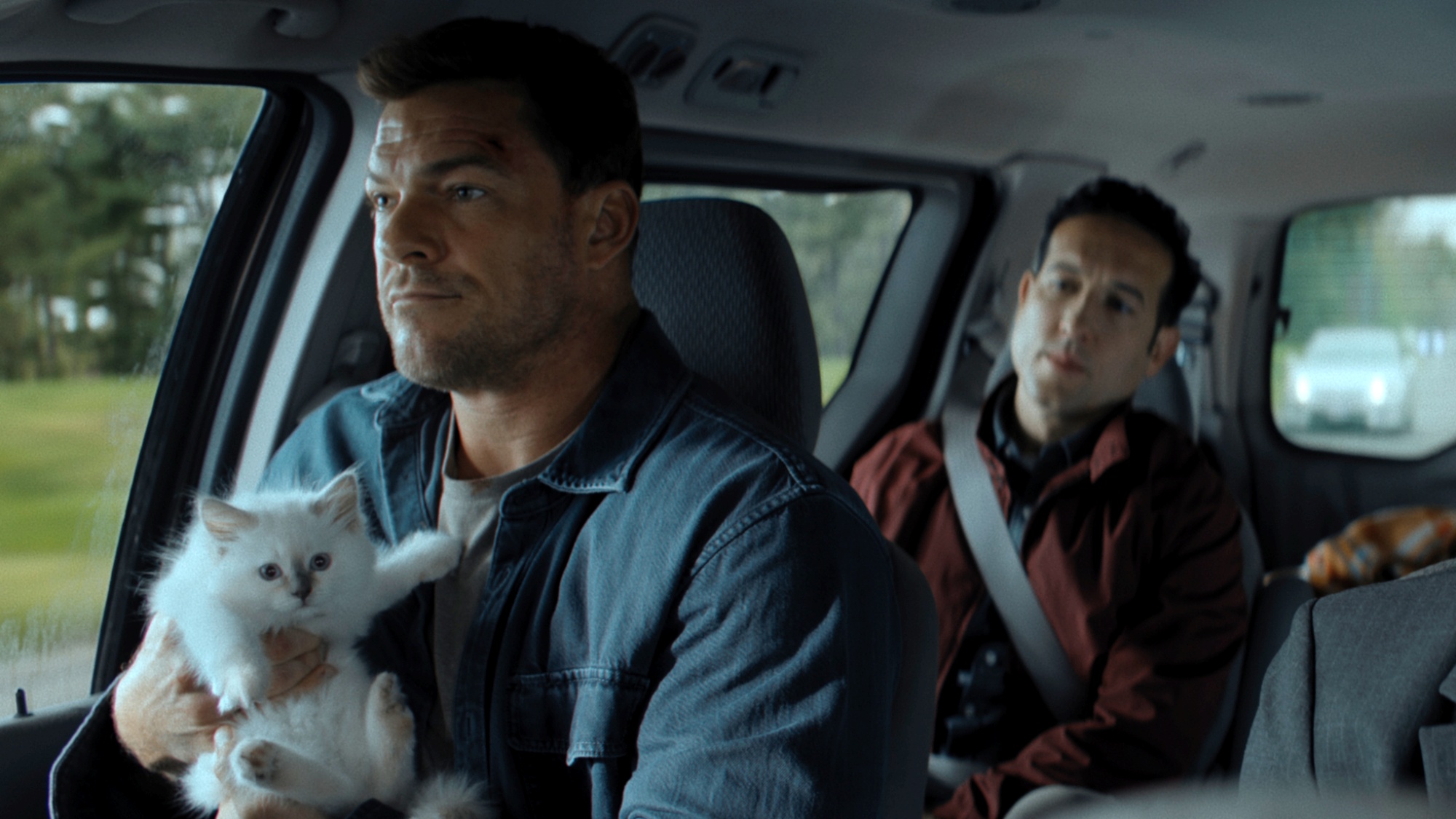 Alan Ritchson as Jack Reacher sitting in the passenger seat of a car while holding a white kitten in Reacher Season 4.