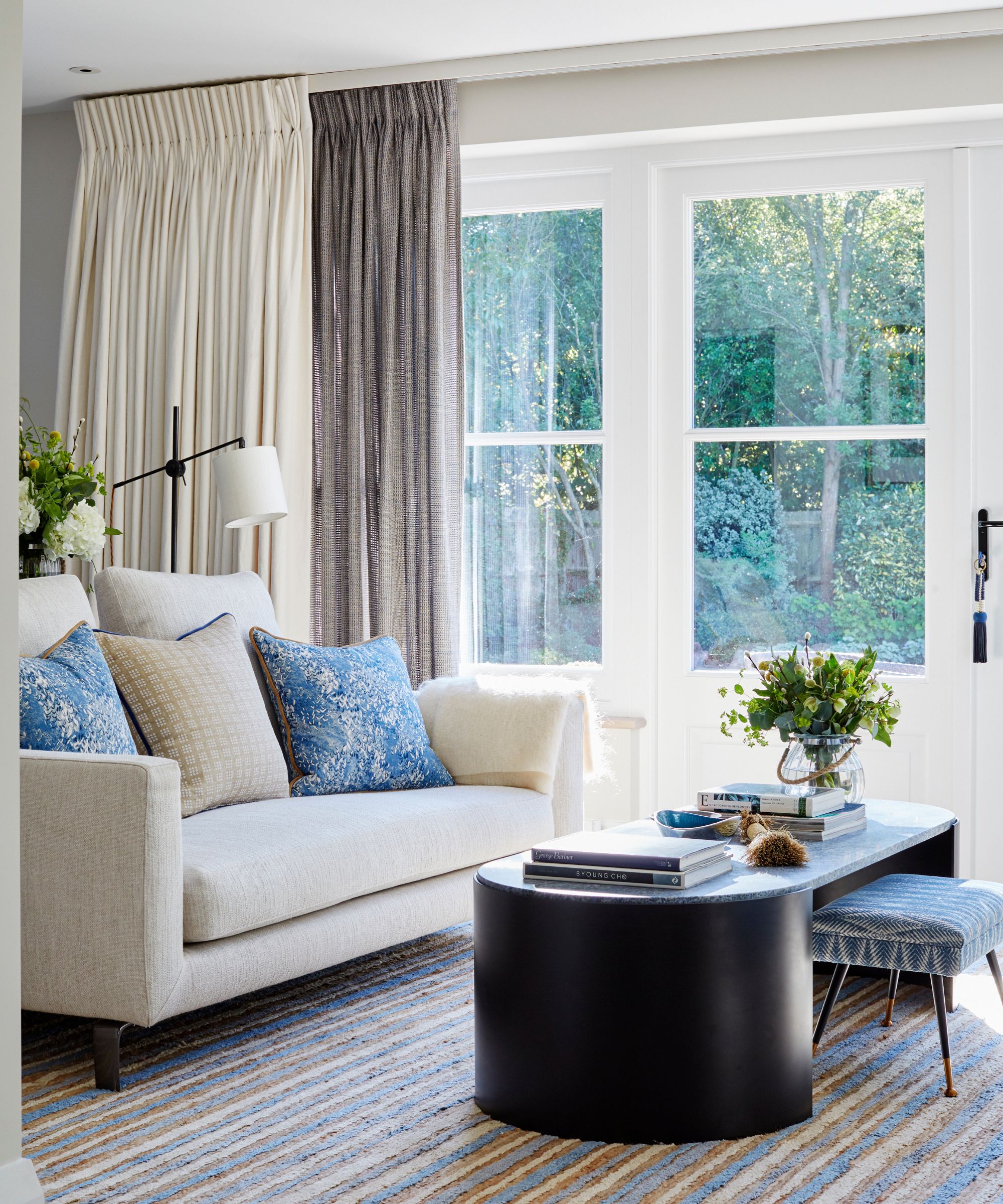 A bright living room with striped rug, black coffee table with flowers and coffee table books, white couch with cushions, white and black floor lamp, and large doors to a green leafy garden with a double layer of grey and white curtains.