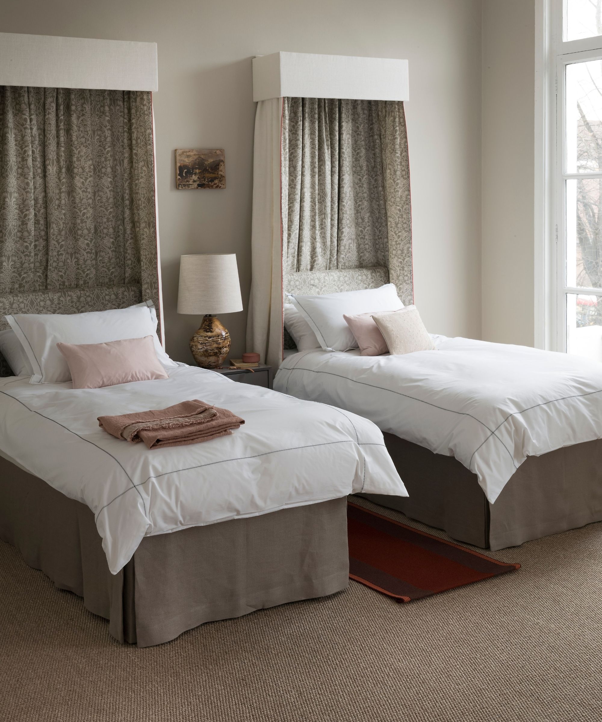 Neutral twin bedroom with matching bed canopies, white bedding, and jute carpeting, with a large window to the right.