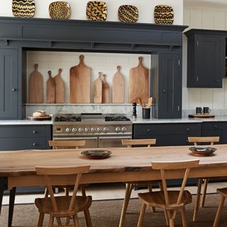 Kitchen with a long wooden table with chairs, grey cupboards and wooden chopping boards hanging on the wall