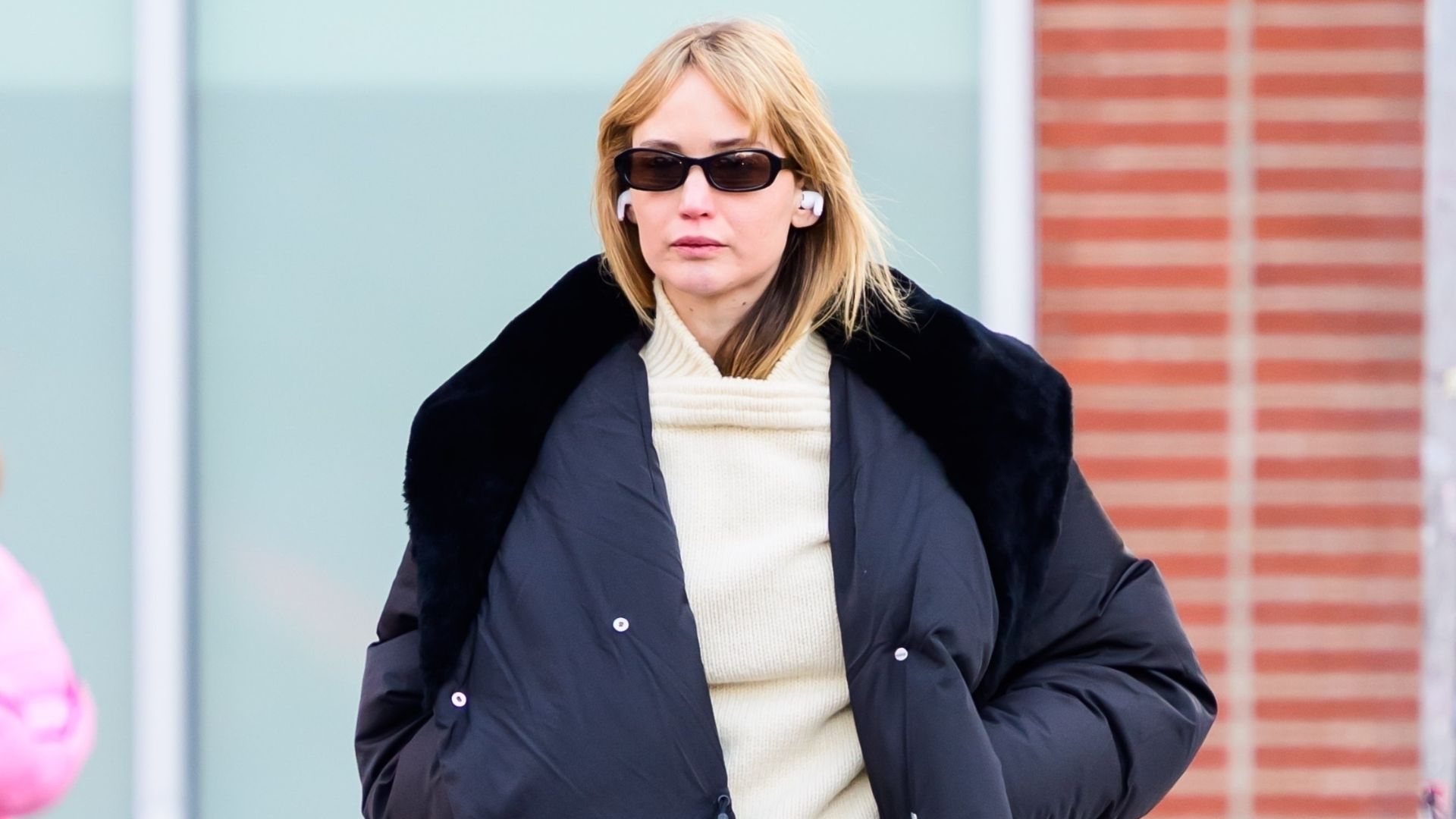 Jennifer Lawrence walks down the street in New York wearing a puffer jacket with a butter yellow jumper and a pink scarf tied around her waist.