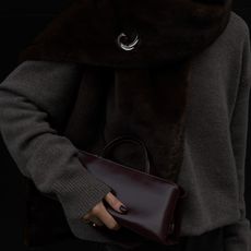 Woman wears a grey sweater, fur stole with silver pin and Verafied's eclair bag in burgundy.