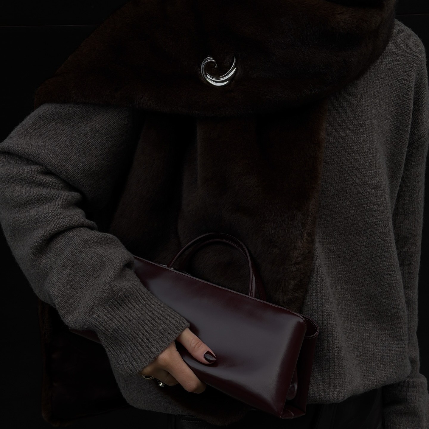 Woman wears a grey sweater, fur stole with silver pin and Verafied's eclair bag in burgundy.