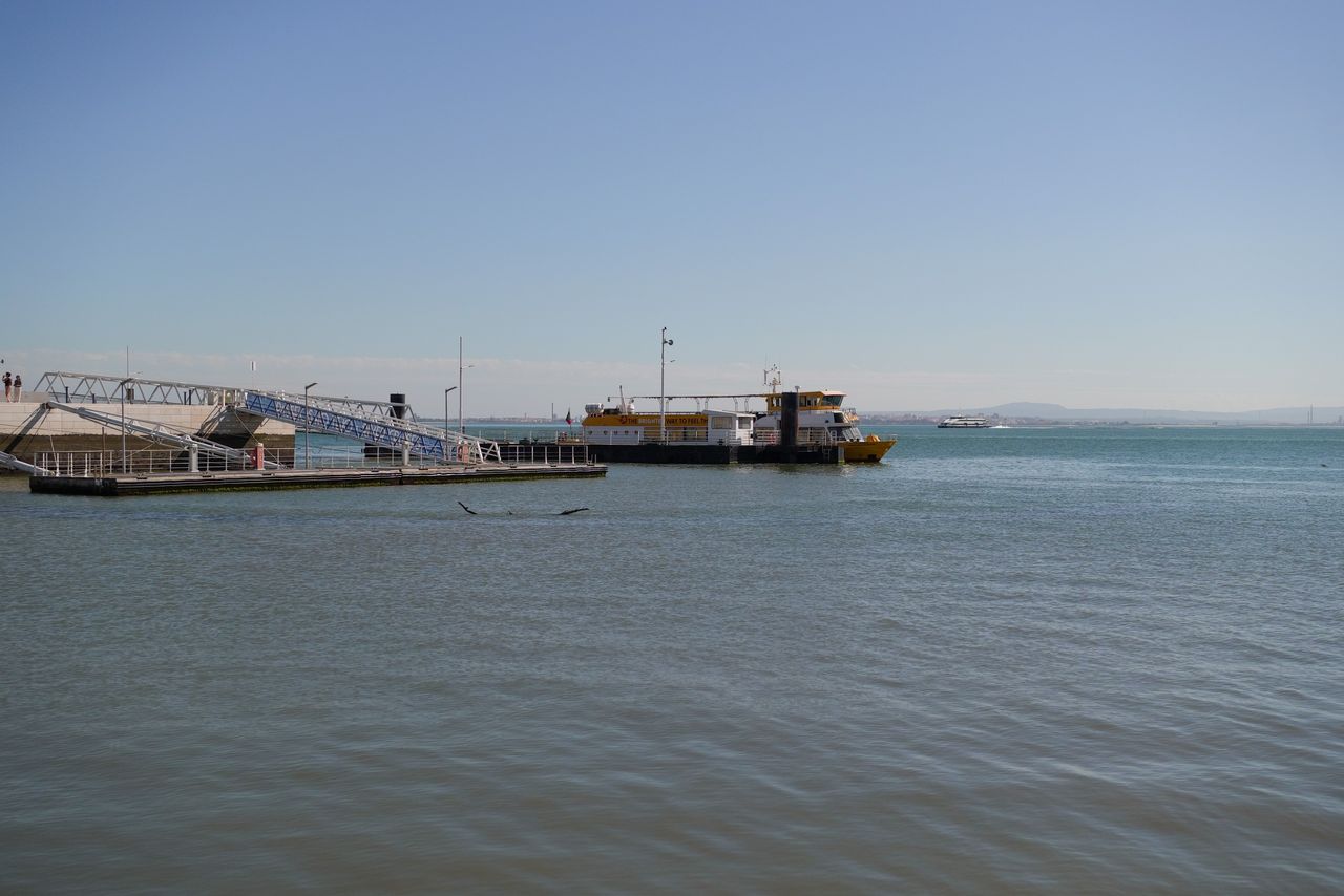 Ferry moored beside a riverside terminal in Lisbon, with calm water and distant shoreline beneath a clear sky.