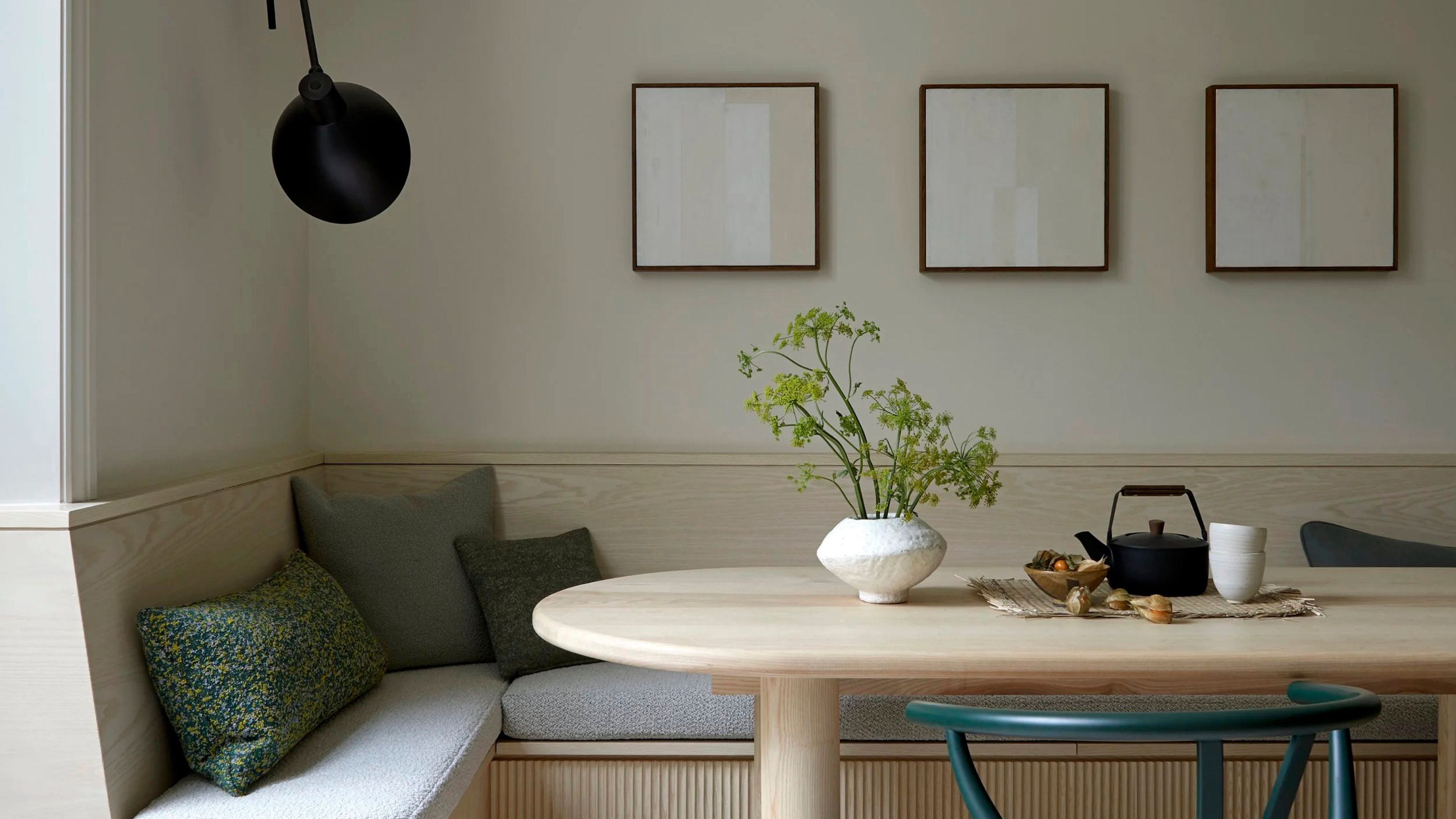 calm dining room with built-in timber banquette seat with grey cushion and sloped timber backrest, cream walls styled with three artworks and a mounted wall lamp, a timber table styled with a vase, teapot and mugs, and a green armchair