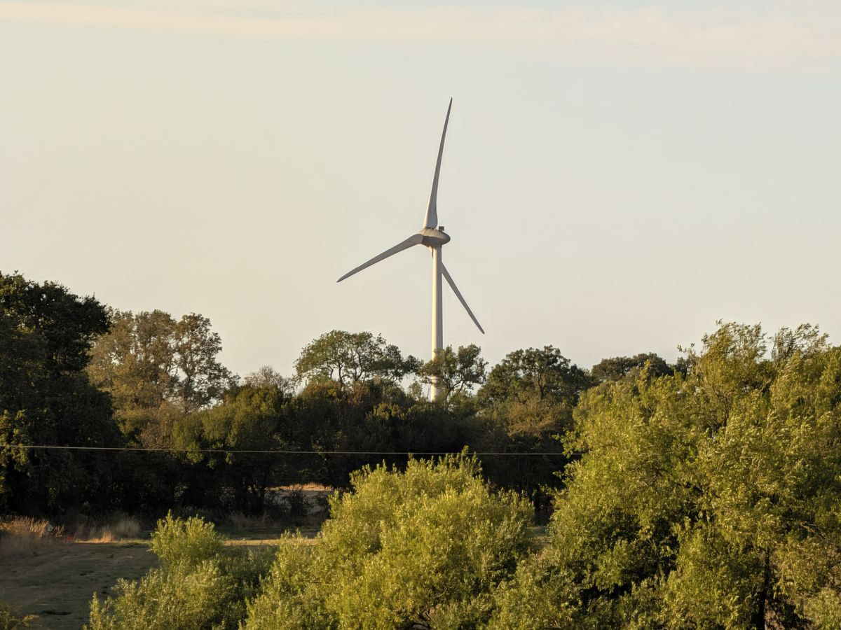 Wind turbine rising above trees and dry fields in a rural landscape lit by warm evening sunlight.