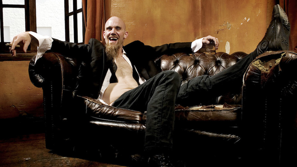 Nick Oliveri sprawled on an old sofa, holding a cigar and a glass of whiskey.