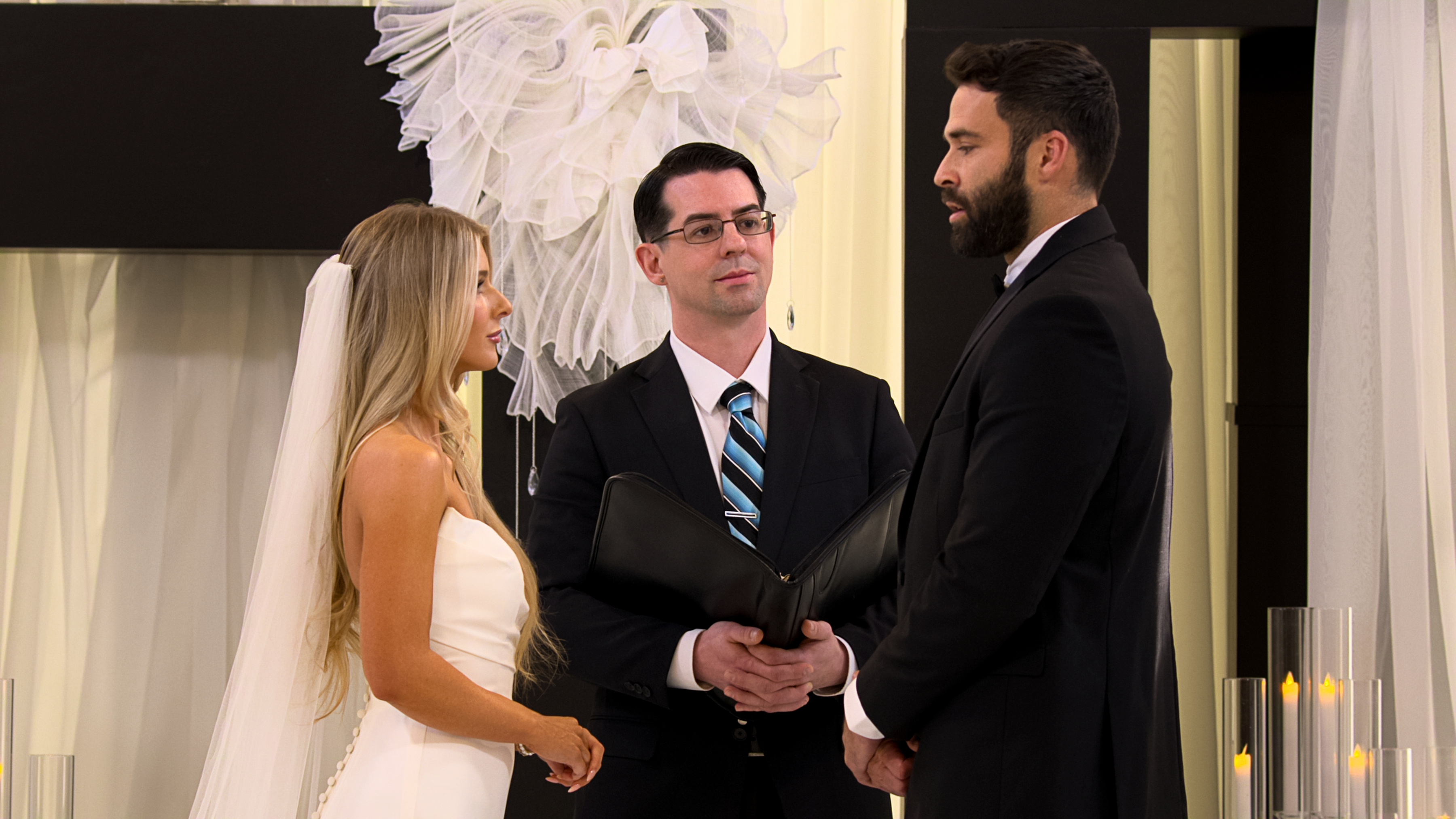 A woman in a wedding dress and veil (Ashley Carpenter, left) stands with a man in a tux (Alex Henderson, right) at the altar, in front of a male officiant (center) in the 'Love Is Blind' season 10 finale.