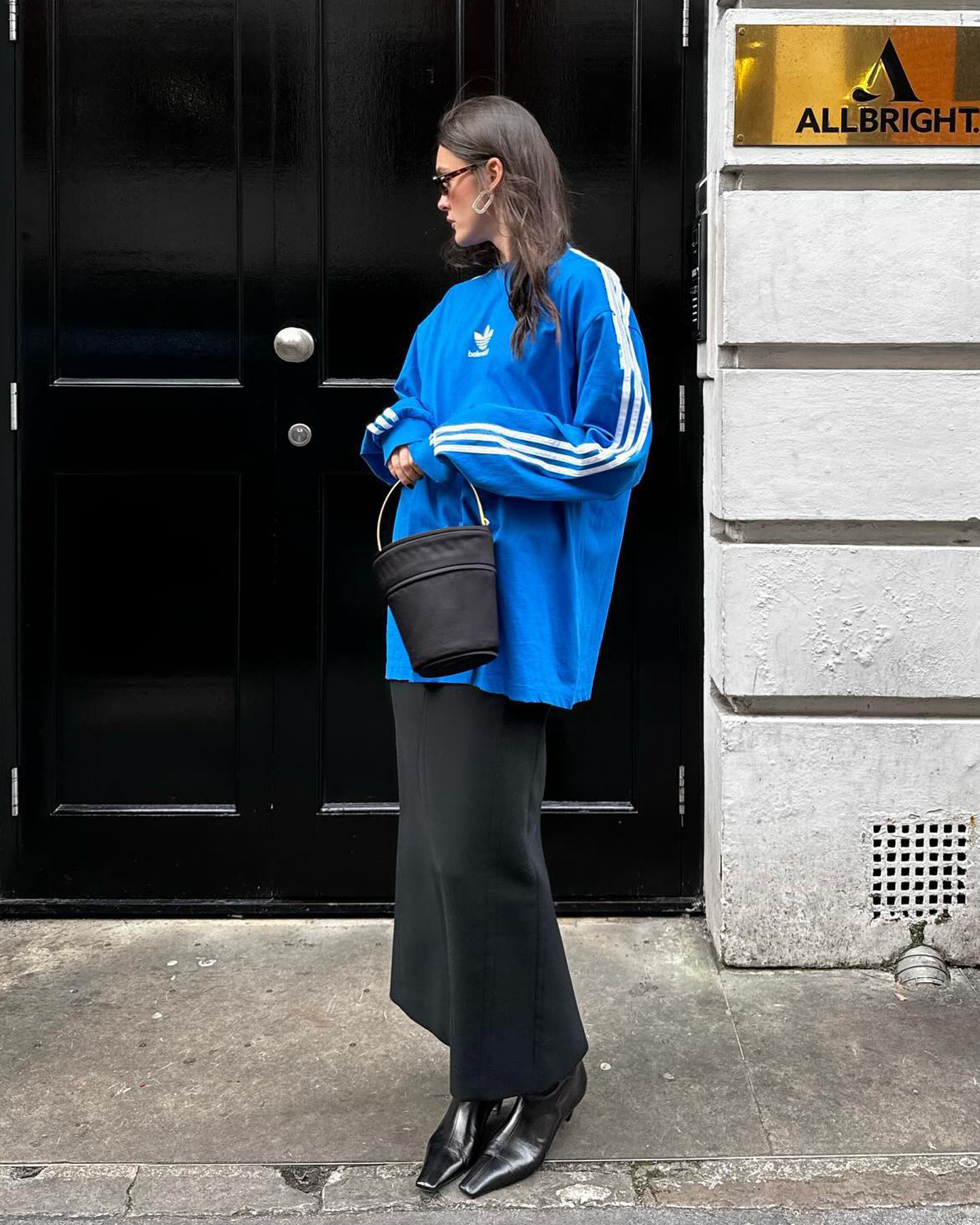 fashion influencer poses on the sidewalk in London wearing a bright blue Adidas long sleeved top with a black bucket bag, a black maxi skirt, and square-toed boots