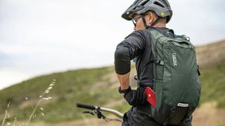 A white male mountain biker stands with his back to the camera and he's wearing a green Camelbak MULE
