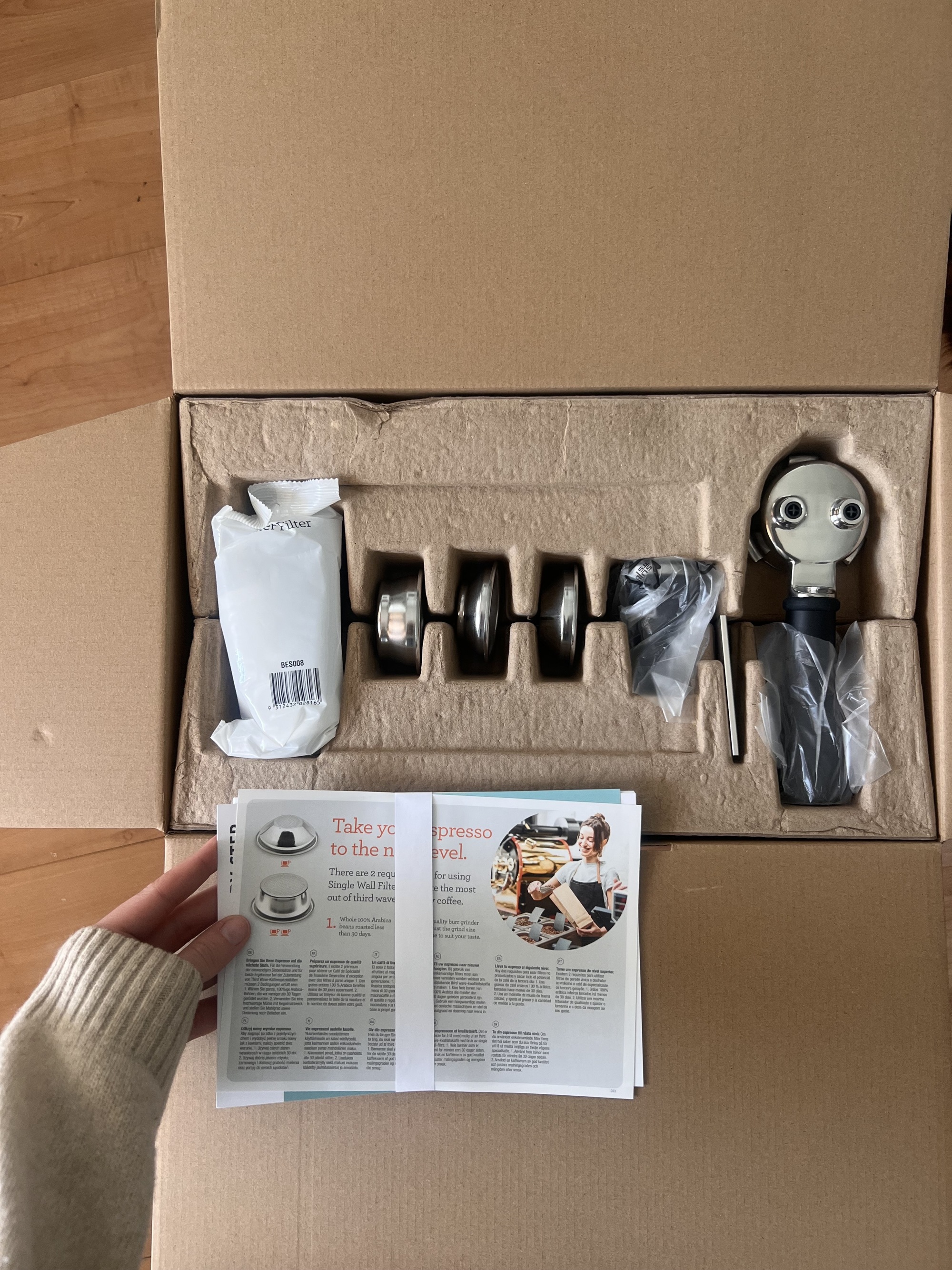 Image of a hand in a beige sweater holding the instruction manual over a cardboard box that has coffee machine accessories  in it.