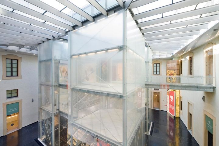 Black gloss stone courtyard floor, white walls with neutral colour doors and black framed windows along the ground and first floor levels, steel and glass towers are positioned in the middle of the courtyard with an opaque covering which blurs the contents inside the towers, girder and windowed roof letting light into the space below