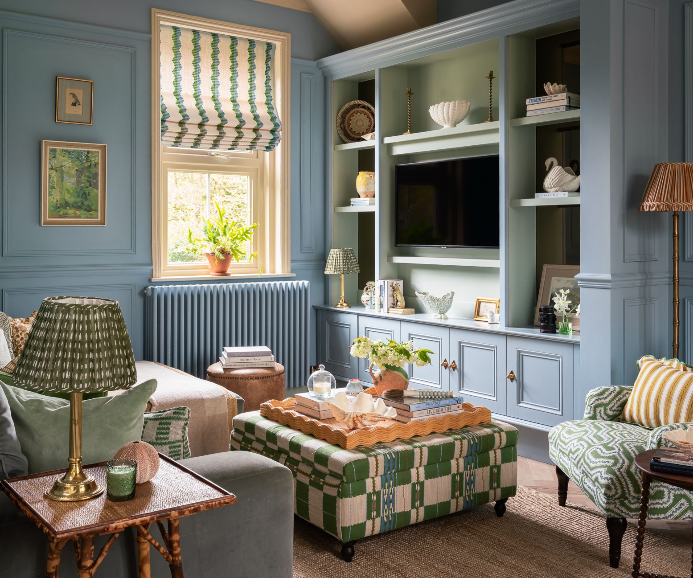 Powder blue living room with built-in bookcase housing TV, green geometric ottoman, blue sofa with green cushions, and green armchair with stripey yellow cushion