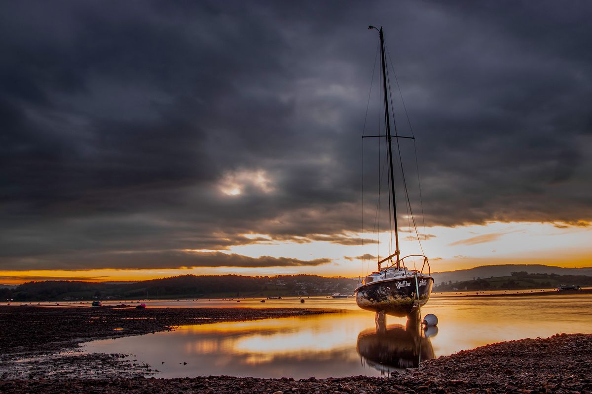 Sailing boat at low tide in front of moody sky with bright sunrise