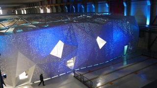 A photo of the Lumi supercomputer in Kajaani, Findland, shot from a gantry overlooking a large hangar containing the supercomputer housing. The housing is angular and lit in blue, with holes throughout giving it the appearance of stars in an evening sky. A person walks in front of the housing to the bottom left of frame and is dwarfed by the structure.