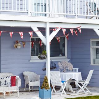 blue porch with balcony above, white rattan bench seating with wood table and chairs, bunting and planters