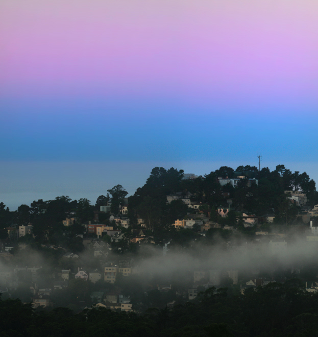 Layers of purple and blue and gray are seen in a clear sky above a hilly city full of trees