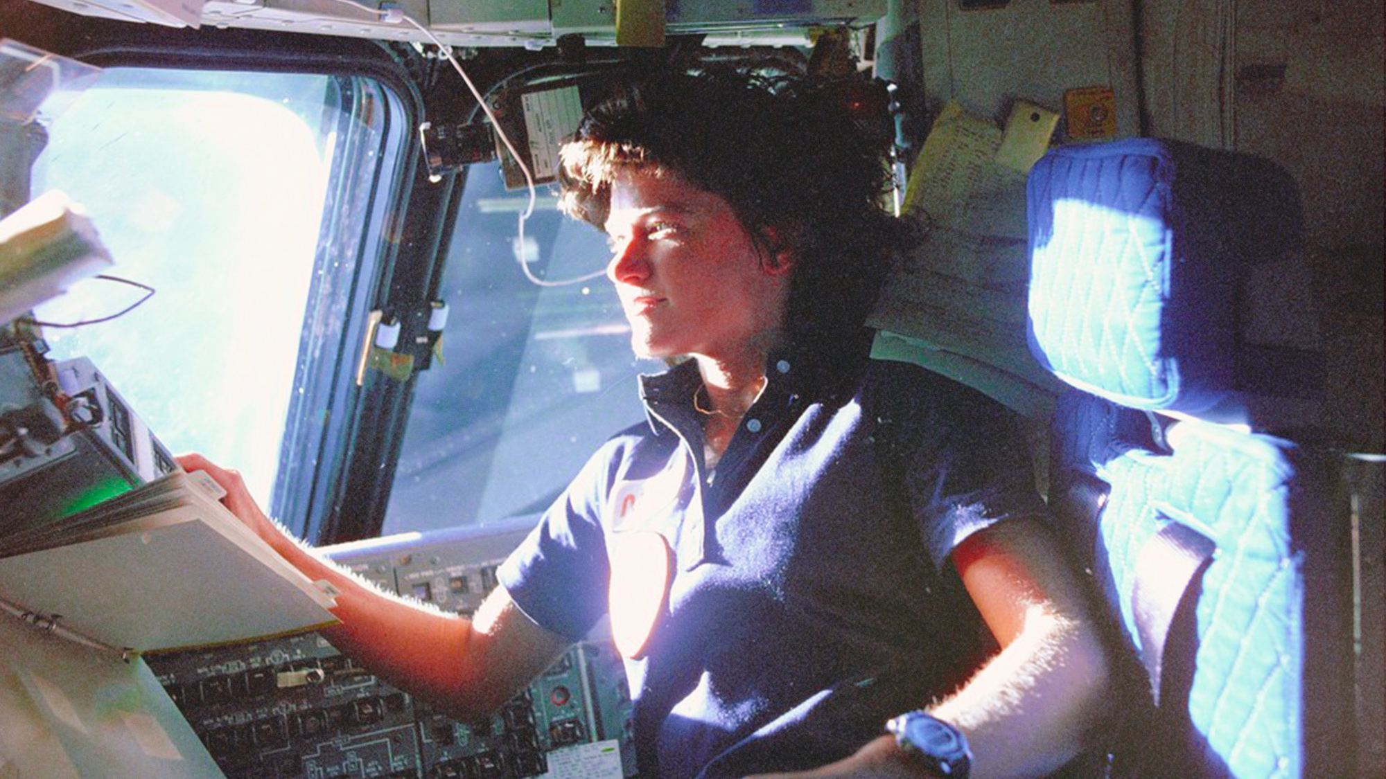 Astronaut Sally Ride, mission specialist on STS-7, monitors control panels from the pilot’s chair on the Flight Deck. Floating in front of her is a flight procedures notebook