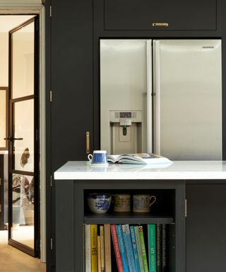 A kitchen with a black kitchen island with colorful books and mugs in the shelves, a white counter with a blue mug and cooking book on top, and a silver fridge, black cabinets, and a glass door behind it