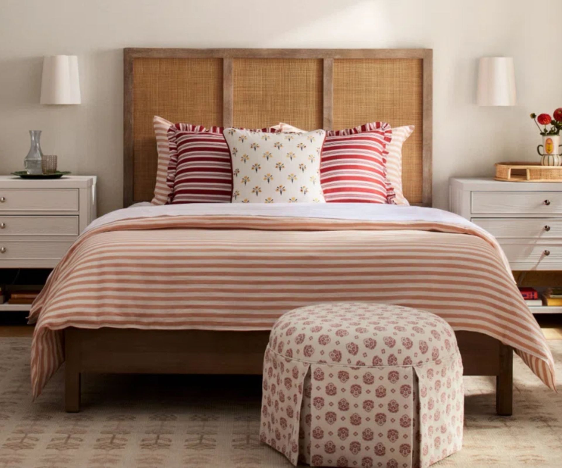 pink bedroom with a rattan and wooden bed, pink striped bedding a pink block print upholstered ottoman at the foot of the bed