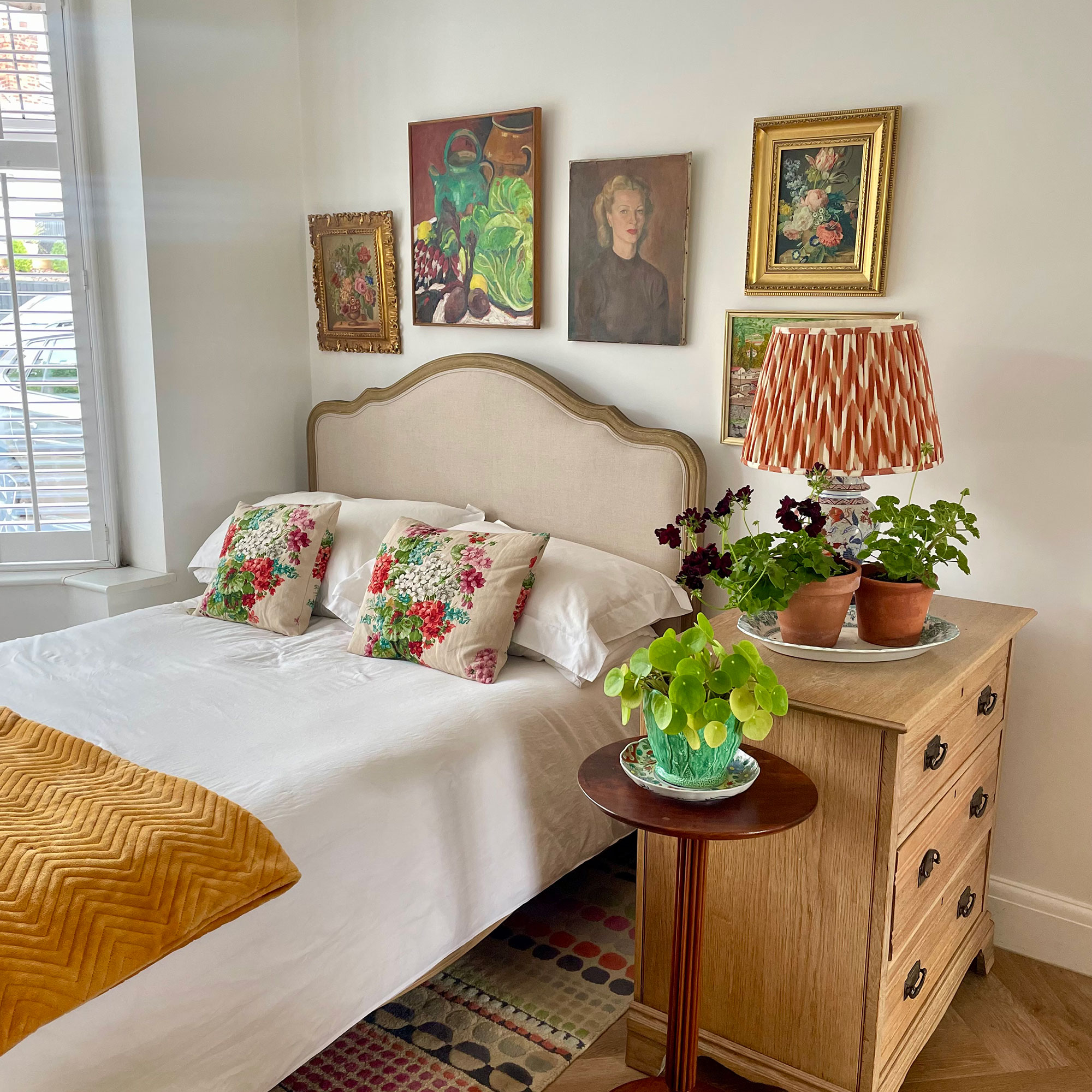 Bedroom area with gallery wall above bed, with bedside table holding table lamp and houseplants