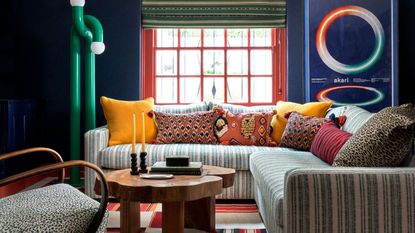 A living room with dark blue walls, red window trim, a green striped corner sofa, a wooden coffee table, a red and cream checkered rug and an abstract and minimal green lamp. 