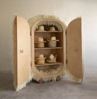 An open cabinet by Fernando Laposse containing shelves displaying straw hats, and doors covered in white fur