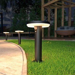 Three black illuminated stake lights in green grass along the edge of a paved pathway