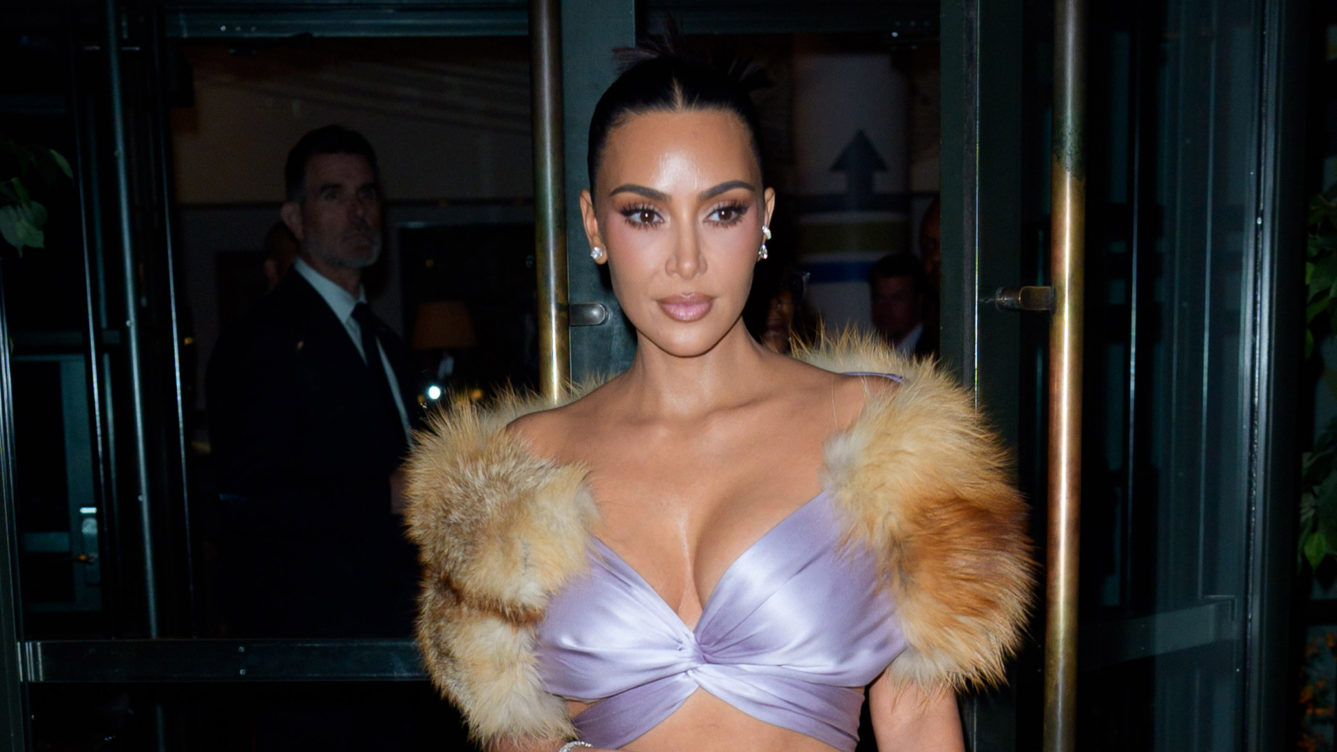 Kim Kardashian wearing a sleek updo hairstyle. She is wearing a top that has fur sleeves and a purple bralette.