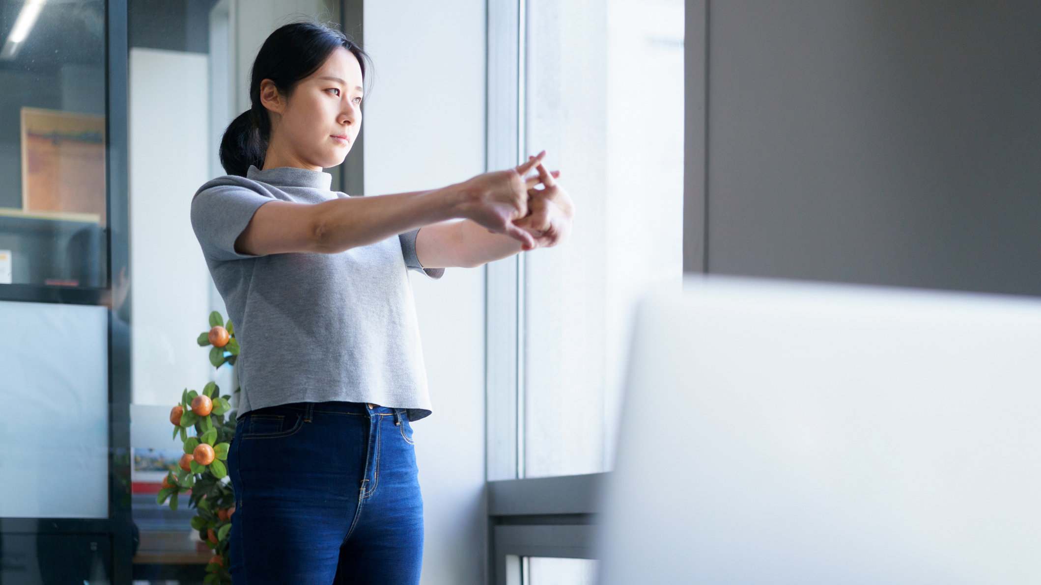 Woman in an office stands and looks out of a window and stretches her wrists, with her fingers interlocked, pushing her palms away from her