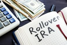"Rollover IRA" handwritten in a notepad on a desk with a calculator, pen and U.S. money nearby