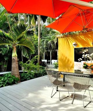 A residential backyard with a yellow outdoor room, a white wooden deck, a glass dining table with rattan chairs, two red canopies above them, and tall leafy trees and shrubbery