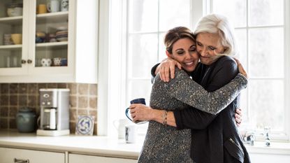 An adult daughter hugs her mother in the kitchen.