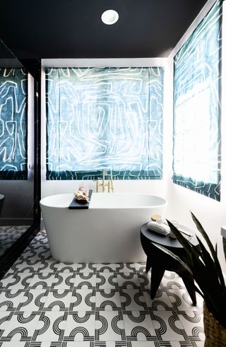 An example of dark bathroom ideas showing a black bathroom with a white bath, black and white floor and blue patterned blinds