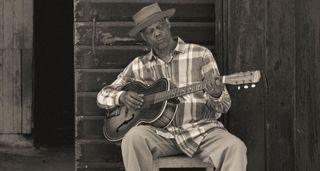 A black-and-white press shot of Eric Bibb playing some acoustic blues on a chair outdoors.